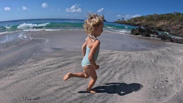 Side view of energetic girl running on wet sand along ocean shore. Wide shot of blonde curly hair girl running to big stones past raging ocean in background Running is great sport on vacation by ocean