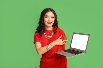 Beautiful Indian woman in sari pointing at laptop on green background