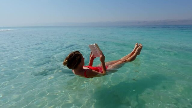 Peaceful Young Woman Floating in the Dead Sea wearing Red Swimsuit and Reading a Book