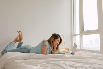 Happy Asian Thai woman using laptop lying on white bed at home, working as freelancer and spending...