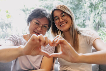 Happy Asian Thai older mother and daughter showing love gesture, smiling making heart sign and looking at camera, sitting on sofa together at home.