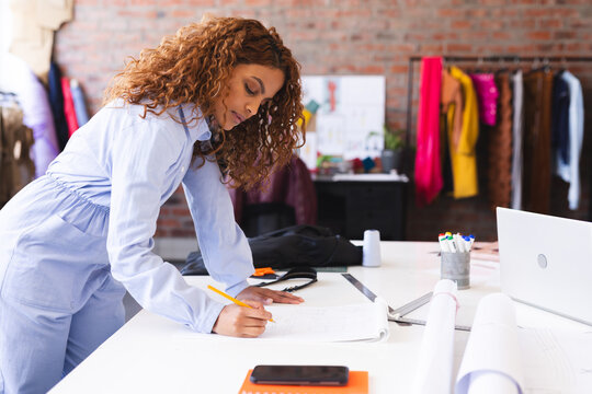 Happy biracial female fashion designer making notes and using smartphone in sunny studio