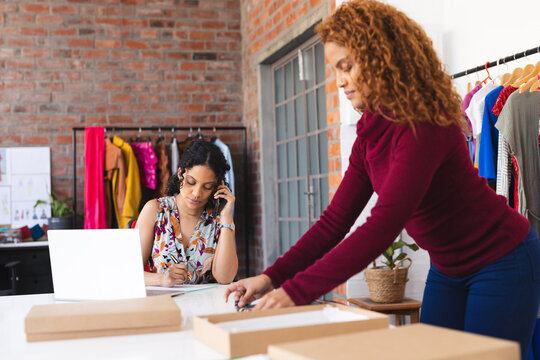 Two biracial female fashion designers packing parcels, using smartphone and laptop in sunny studio