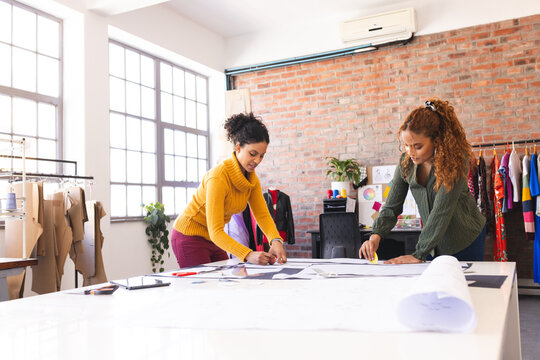 Two happy biracial female fashion designers working with fabric in sunny studio