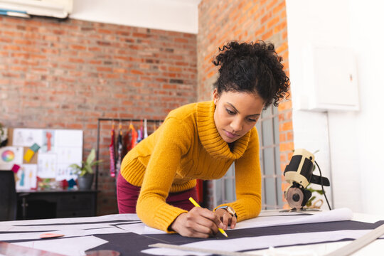 Focused biracial female fashion designer using tailor's pencil in sunny studio