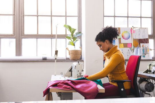 Focused biracial female fashion designer using sewing machine in sunny studio