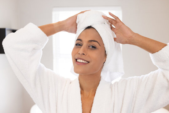 Portrait of happy biracial woman wearing white bathrobe and towel on head in bathroom