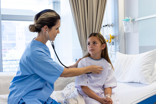 Focused caucasian female doctor using stethoscope on chest of girl patient sitting on hospital bed