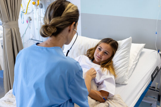 Caucasian female doctor using stethoscope on chest of smiling girl patient lying in hospital bed
