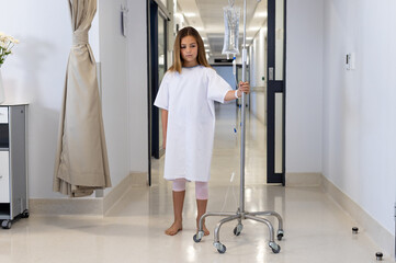 Caucasian girl patient in gown walking with drip stand in hospital corridor