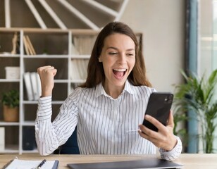 Euphoric young female worker holding mobile phone celebrating win receiving good news about job...