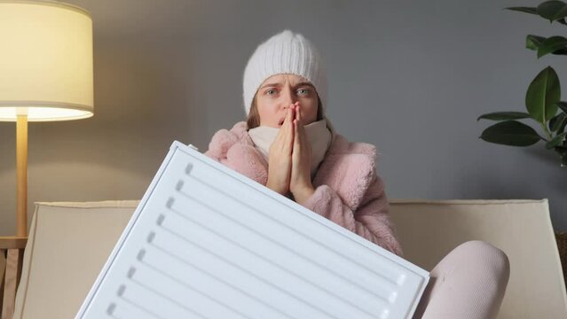 Adapting to seasonal temperature changes and seeking warmth during winter distressed woman bundled up in cap and coat sits on couch clutching heater battery trying to get warm.