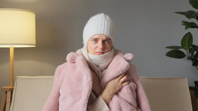 Sad woman have cold on the sofa at home with winter coat and cap suffering from lack of heat looking at camera trying to get warm expressing negative emotions.