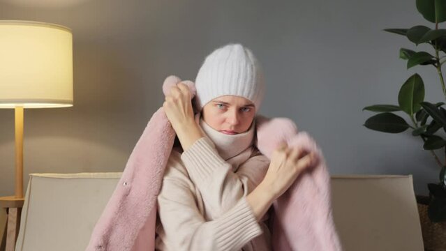 Unwell woman in coat and cap sitting in cold living room suffering from lack of heat at home unhealthy young woman struggling from chill freeze at home.