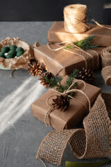 Christmas holiday celebration aesthetic background. Presents, gift boxes wrapped with brown crafted paper, decorated with pine tree cone and twigs, on gray table with natural sunlight