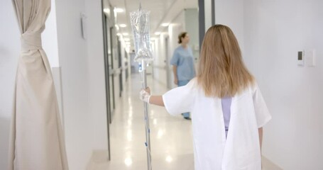 Rear view of caucasian girl patient walking with drip stand in hospital, copy space, slow motion