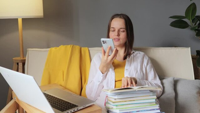 Student woman recording voice message making phone call on speaker talking giving command to electronic voice assistant on smartphone while studying with laptop and books at home.