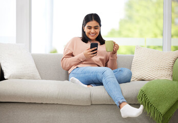 Obraz premium Rest, relaxation and carefree with a young woman typing on a phone, drinking coffee and comfortable on the sofa at home. Relaxed female browsing social media or typing a message with a cup of tea
