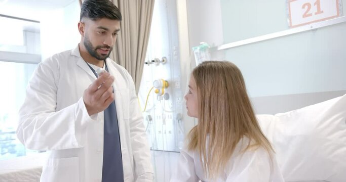 Diverse male doctor showing girl patient in hospital bed how to use asthma inhaler, slow motion