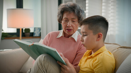 Loving elderly woman retired grandmother reading book with grandson at home on sofa enjoying read...