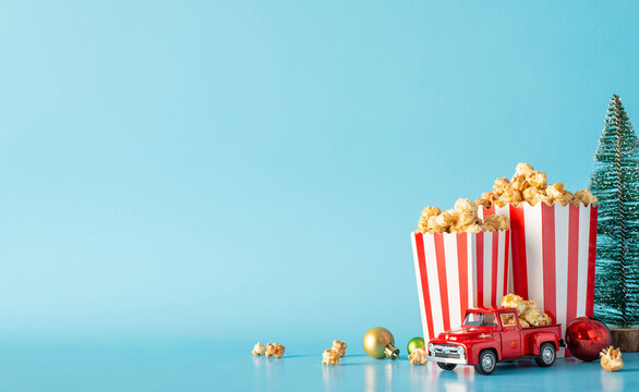 Festive holiday popcorn delivery idea. Side view shot of table adorned with striped popcorn boxes, ornaments, miniature car and a small Christmas tree against a pastel blue wall