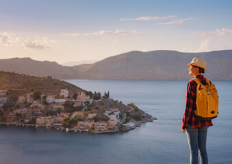Nice asian Happy Female with backpack Enjoying her holidays on Symi Islands in sunset time. View of port Symi or Simi, is tiny island of Dodecanese, Greece, calm atmosphere and fabulous architecture.
