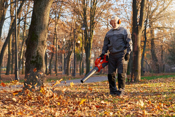 Municipal caucasian worker in age cleaning lawn by blowing out leaf piles of dried leaves outside....