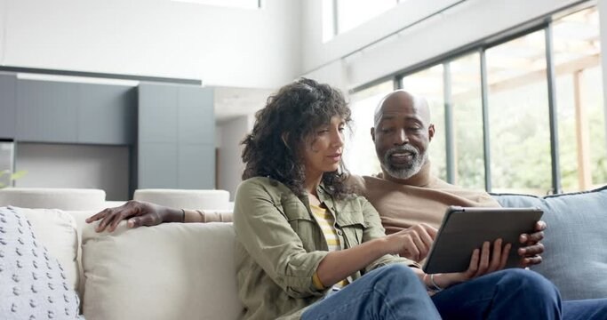 Happy biracial couple sitting on couch and using tablet at home, slow motion