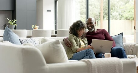 Happy biracial couple sitting on couch and using laptop at home, slow motion - Powered by Adobe