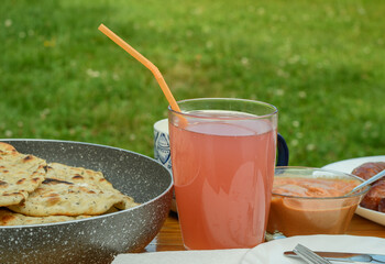 Różowy napój w szklance ze słomką na stole wypełnionym jedzeniem, piknik na trawie - obrazy, fototapety, plakaty