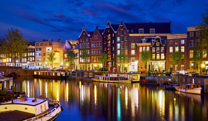 Evening town Amsterdam in Netherlands on bank river canal Amstel with shining window. Panorama landscape brown house over water reflection.