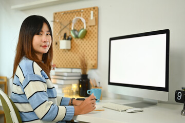 Obraz na płótnie Canvas Portrait of young Asian woman sitting in front of computer monitor and smiling at camera