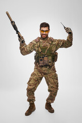 Happy military infantryman jumping with raised arms and rejoicing in studio. Front view of cheerful male soldier with weapon and walkie-talkie dancing, isolated on white. Concept of army, emotions.