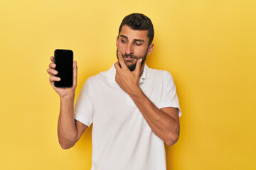 Young Hispanic man shows phone screen looking sideways with doubtful and skeptical expression.