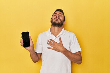 Young Hispanic man shows phone screen laughs out loudly keeping hand on chest.