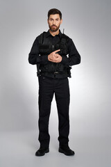 Attractive male cop with ammunition holding gun with both hands. Front view of bearded man in black body armor looking at camera, on gray studio background. Concept of work, police, rescue.