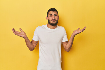Young Hispanic man on yellow background doubting and shrugging shoulders in questioning gesture.