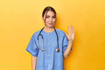 Young nurse in a yellow studio smiling cheerful showing number five with fingers.