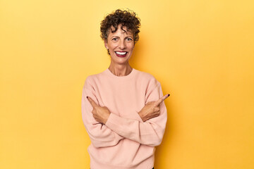 Mid-aged caucasian woman on vibrant yellow points sideways, is trying to choose between two options.