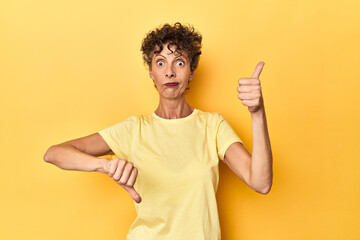 Mid-aged caucasian woman on vibrant yellow showing thumbs up and thumbs down, difficult choose...