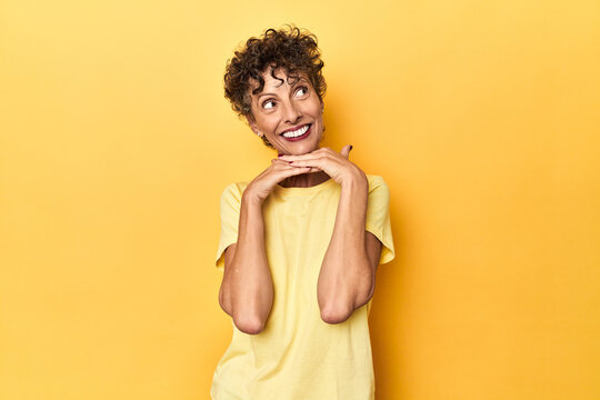 Mid-aged caucasian woman on vibrant yellow keeps hands under chin, is looking happily aside.