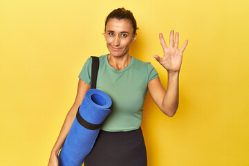 Middle aged sportswoman with yoga mat on yellow studio smiling cheerful showing number five with...