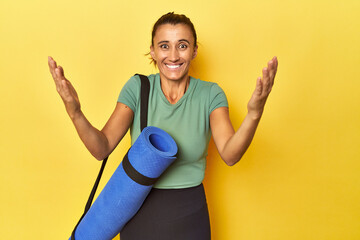 Middle aged sportswoman with yoga mat on yellow studio receiving a pleasant surprise, excited and...