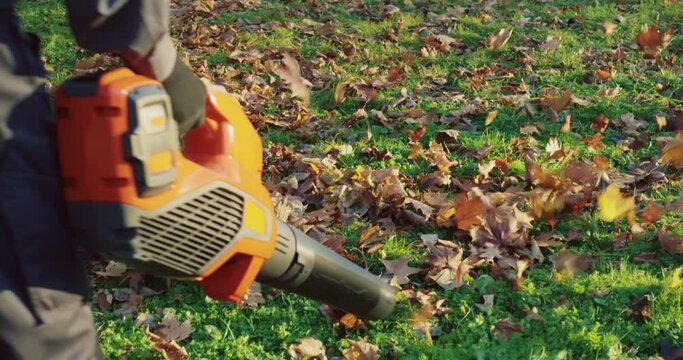 Close up of caucasian concierge in working uniform removing dry fall leaves at city park with hand blower. Concept of people, landscaping and maintaining.