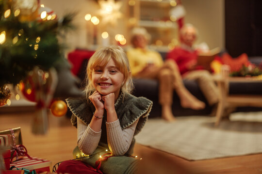 Little happy girl packing Christmas present in front of a Christmas tree and smiling at camera