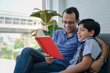 Happy indian father reading book with his son in living room.
