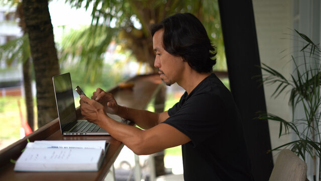 Asian person use mobile cell phone outside palm tree cafe place. Busy freelancer sit work outdoor. Smart people write macbook. Man chat iphone user. Freelance home job. Male guy boss study nature Asia