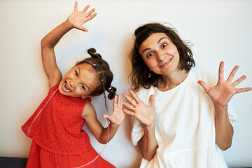 Babysitter and little girl in red dress having fun playing together at home against white wall, grimacing to frighten you, child making scary face, woman looking at camera with big eyes and smile - obrazy, fototapety, plakaty