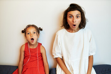 Shocked surprised young female and little girl looking at camera with open mouth and big eyes, astonished with jaw dropping news, sitting on back of couch against white wall at home - Powered by Adobe