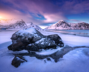 Sandy arctic beach with blue sea, stone in frozen coast and rocks in snow in winter at sunset in Lofoten islands, Norway. Colorful Landscape with snowy mountains, sky with clouds. Norwegian coast - obrazy, fototapety, plakaty